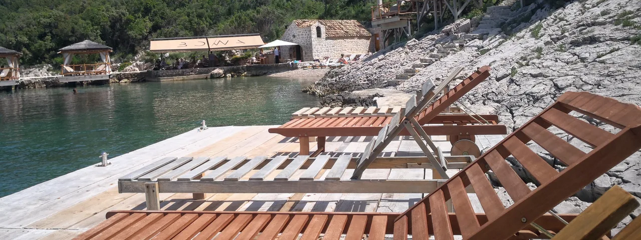 Tranquil bay with wooden lounge chairs on rocky shore, overlooking crystal-clear waters and lush green hillside in Elafiti Islands