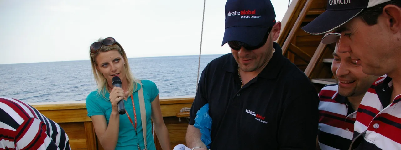 A group participates in a team-building activity on a ship deck in Dubrovnik. A woman with a microphone gives instructions as team members work together with ropes.