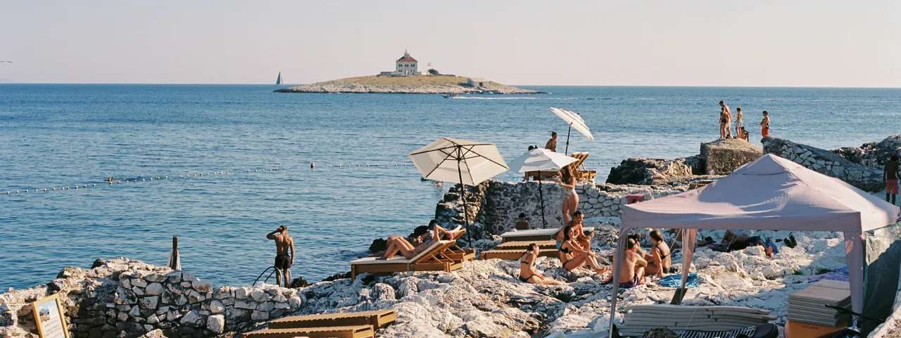 People sunbathing on a rocky Croatian beach with umbrellas, enjoying the Adriatic Sea view and a distant island under clear skies.