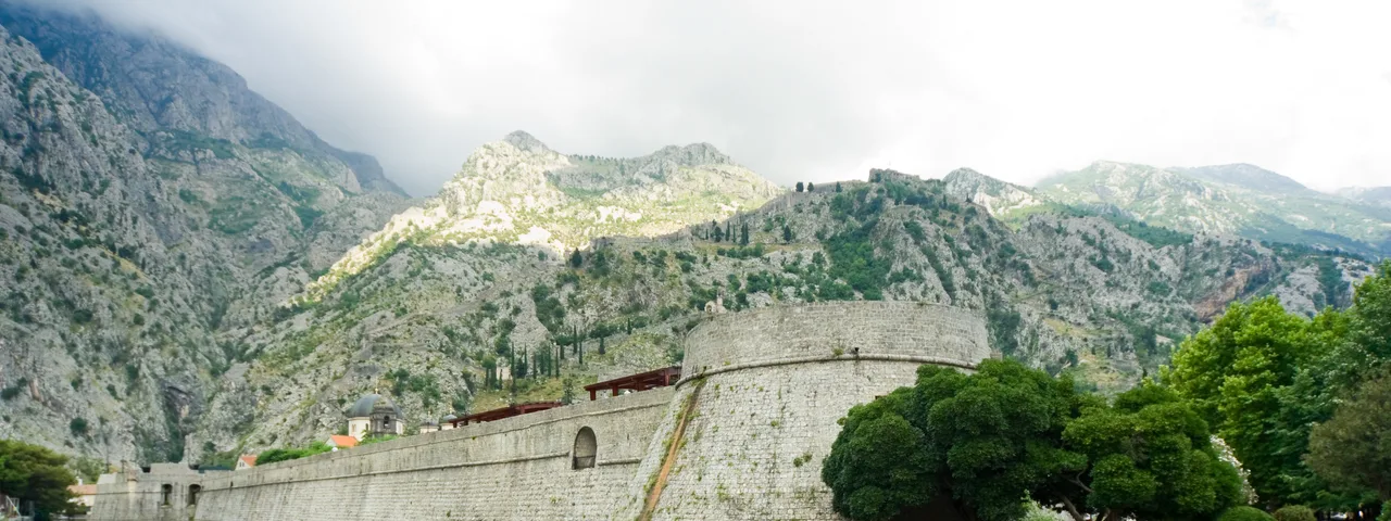 Massive medieval city walls of Kotor reflected in still green moat water with karst mountains behind