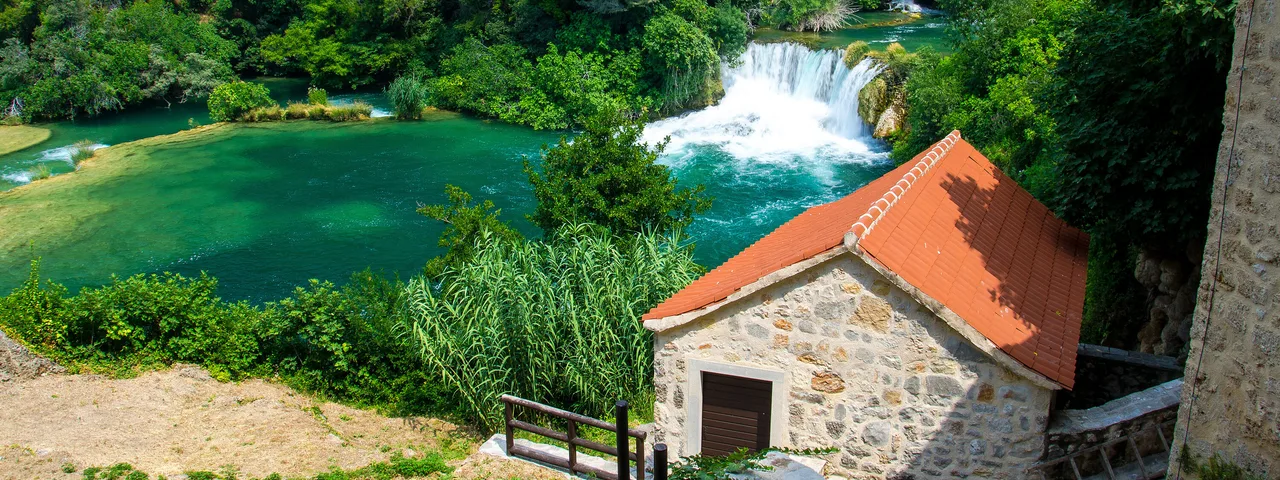 Historic stone mill with terracotta roof beside a waterfall cascading into emerald green pool at Skradinski Buk, Krka National Park, Croatia