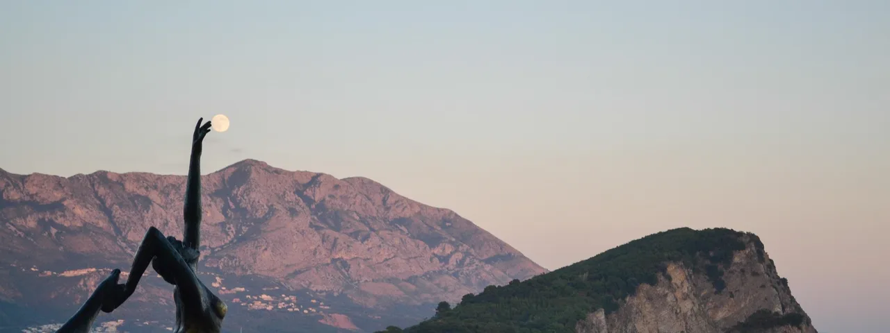 Budva's Ballerina (Dancer of Budva) bronze statue on the rocks at dusk with the full moon over Lovćen mountain and Sveti Nikola island in the background, Montenegro
