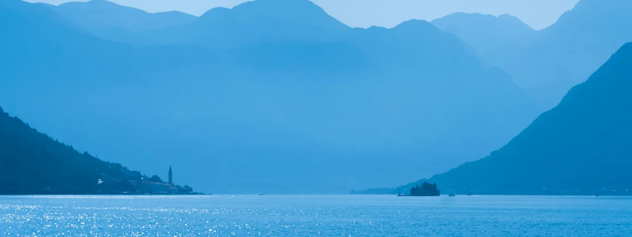 Layered misty blue mountains over the Bay of Kotor with the bell tower of Perast visible on the shoreline, Montenegro