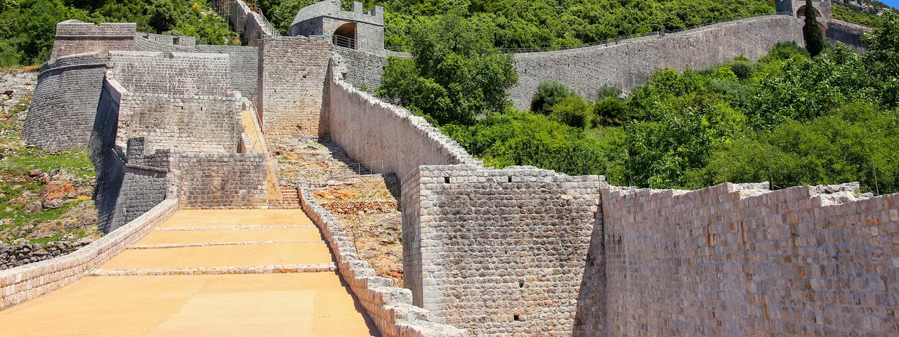 Ston defensive walls zigzagging steeply up a rocky green hillside with worn stone walkway in the foreground, Croatia