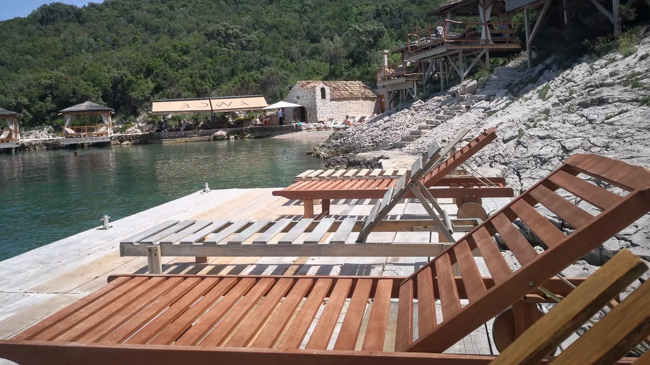 Tranquil bay with wooden lounge chairs on rocky shore, overlooking crystal-clear waters and lush green hillside in Elafiti Islands