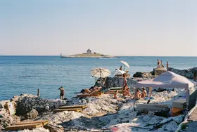 People sunbathing on a rocky Croatian beach with umbrellas, enjoying the Adriatic Sea view and a distant island under clear skies.
