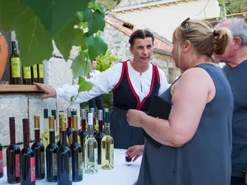 A woman in traditional Croatian dress presents a selection of locally produced red and white wines to visitors. Dried prosciutto hangs in the background, along with grapevines.