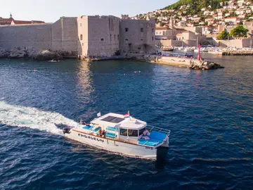 Catamaran leaving a vibrant wake on the deep blue Adriatic Sea near ancient Dubrovnik city walls, Croatia.