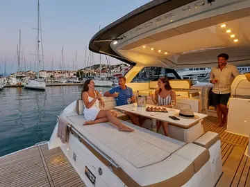 Four people socialize and relax on the deck of a modern yacht, which is moored in a marina filled with various sailboats and vessels under a soft evening sky.