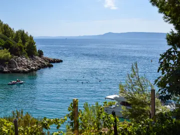 View of a clear blue bay with a small boat and yacht, rocky coastline, and lush greenery from a vineyard overlooking the Adriatic Sea near Split, Croatia.