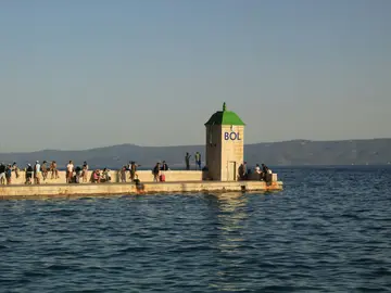 View of Bol pier on Brac island, Croatia, with people gathered around a small building by the calm Adriatic Sea and mountains.