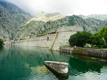 Massive medieval city walls of Kotor reflected in still green moat water with karst mountains behind