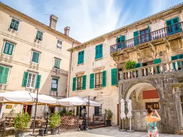 Tourist photographing historic stone buildings and green shutters of Kotor old town square, Montenegro