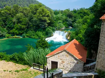 Historic stone mill with terracotta roof beside a waterfall cascading into emerald green pool at Skradinski Buk, Krka National Park, Croatia