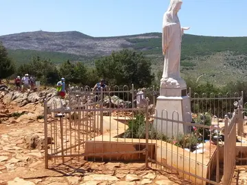 White statue of Our Lady of Medjugorje on Apparition Hill surrounded by iron fence and pilgrims