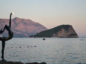 Budva's Ballerina (Dancer of Budva) bronze statue on the rocks at dusk with the full moon over Lovćen mountain and Sveti Nikola island in the background, Montenegro