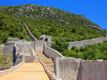 Ston defensive walls zigzagging steeply up a rocky green hillside with worn stone walkway in the foreground, Croatia