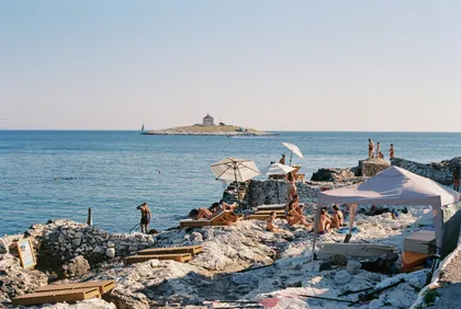 People sunbathing on a rocky Croatian beach with umbrellas, enjoying the Adriatic Sea view and a distant island under clear skies.