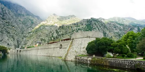 Massive medieval city walls of Kotor reflected in still green moat water with karst mountains behind