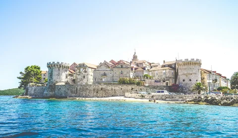 Ancient walled coastal town with stone buildings overlooking crystal-clear turquoise waters of the Adriatic Sea.