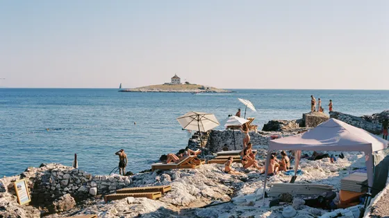 People sunbathing on a rocky Croatian beach with umbrellas, enjoying the Adriatic Sea view and a distant island under clear skies.