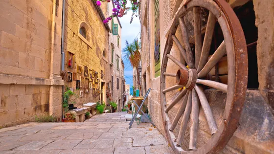 Narrow cobblestone alley in Korčula old town with antique wooden cartwheel, framed paintings and pink bougainvillea overhead