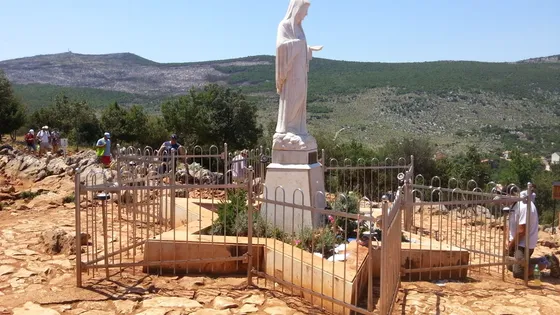 White statue of Our Lady of Medjugorje on Apparition Hill surrounded by iron fence and pilgrims