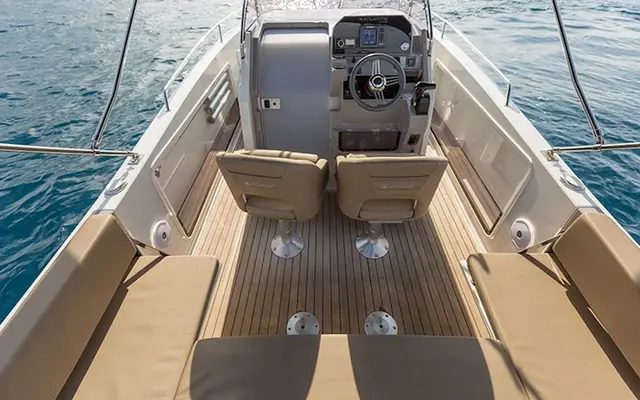 An overhead view of a modern powerboat's deck, featuring tan upholstered seating, wood-grain flooring, and a detailed helm station with a steering wheel, surrounded by blue water.