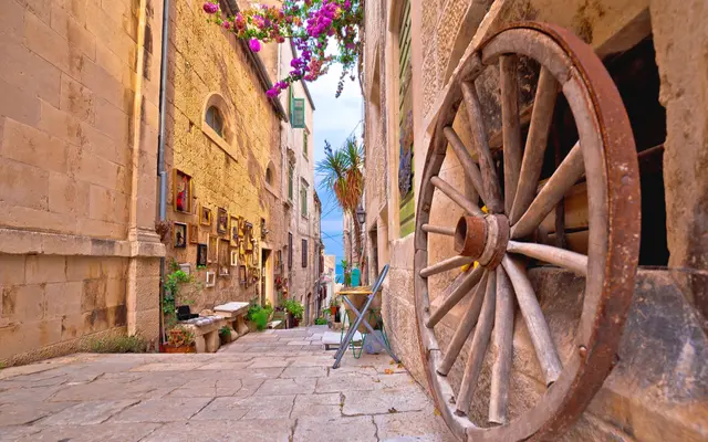 Narrow cobblestone alley in Korčula old town with antique wooden cartwheel, framed paintings and pink bougainvillea overhead