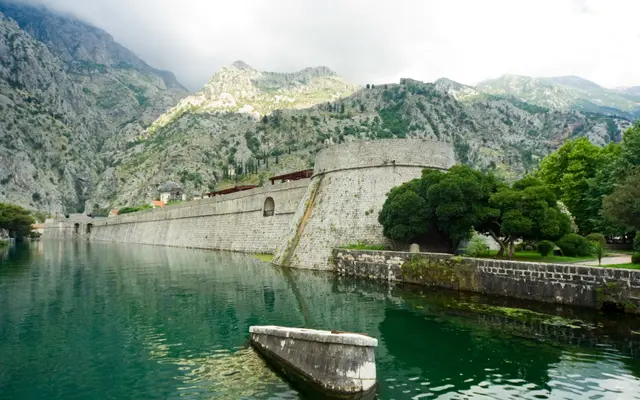 Massive medieval city walls of Kotor reflected in still green moat water with karst mountains behind