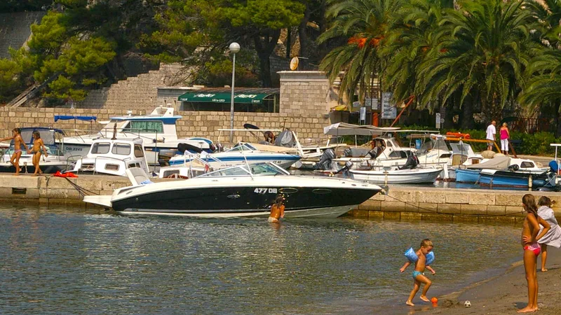 Scenic Dubrovnik Lopud island with various leisure boats, children playing on sunny beach, adults walking along stone harbor, lush Mediterranean coastal vacation scenery, Adriatic Sea activities.