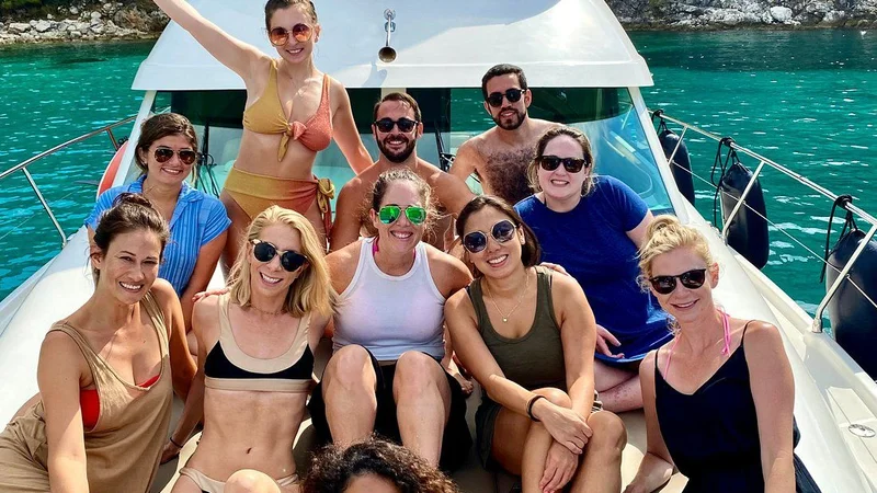 Twelve adults smile for a photo on the deck of a motor yacht, with sparkling turquoise water visible behind them.