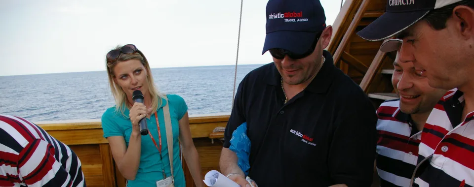 A group participates in a team-building activity on a ship deck in Dubrovnik. A woman with a microphone gives instructions as team members work together with ropes.