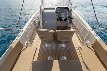 An overhead view of a modern powerboat's deck, featuring tan upholstered seating, wood-grain flooring, and a detailed helm station with a steering wheel, surrounded by blue water.