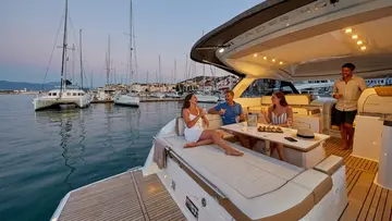 Four people socialize and relax on the deck of a modern yacht, which is moored in a marina filled with various sailboats and vessels under a soft evening sky.