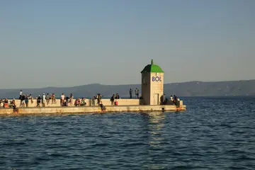 People gather on the scenic pier in Bol, Brač Island, Croatia, with a building marked 'BOL' and calm Adriatic waters.
