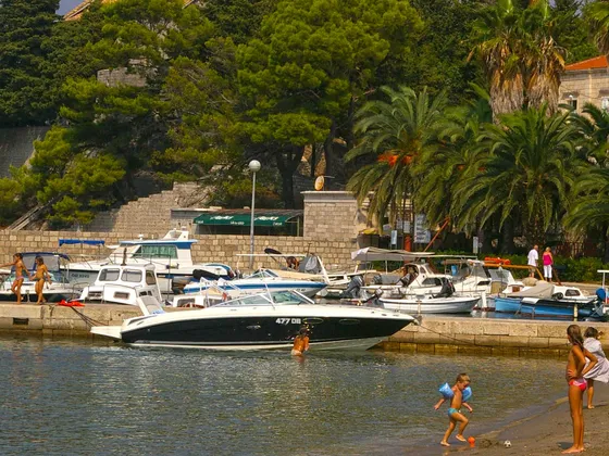 Scenic Dubrovnik Lopud island with various leisure boats, children playing on sunny beach, adults walking along stone harbor, lush Mediterranean coastal vacation scenery, Adriatic Sea activities.