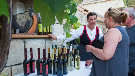 A woman in traditional Croatian dress presents a selection of locally produced red and white wines to visitors.  Dried prosciutto hangs in the background, along with grapevines.