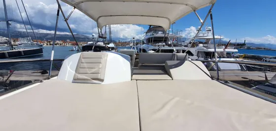 Spacious deck of a luxury yacht featuring plush, light-colored sun pads and a comfortable shaded lounge area. The yacht is moored in a beautiful, bustling marina with other high-end vessels, overlooking a charming coastal town with traditional architecture and majestic mountains under a blue sky. The scene evokes relaxation and upscale travel.