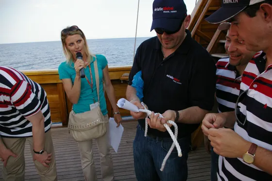 A group participates in a team-building activity on a ship deck in Dubrovnik. A woman with a microphone gives instructions as team members work together with ropes.