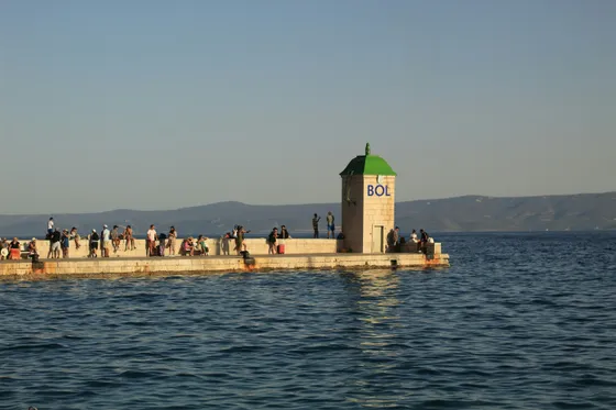 People gather on the scenic pier in Bol, Brač Island, Croatia, with a building marked 'BOL' and calm Adriatic waters.