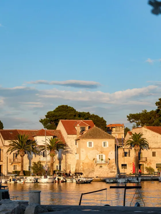 Picturesque Milna village on Brač Island, Croatia, with historic stone buildings, palm trees, and boats docked in its serene harbor at sunset.