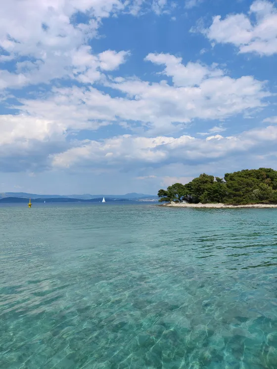 View of crystal-clear turquoise waters of Blue Lagoon near Split, Croatia, with a green island and sailboat under a blue sky.