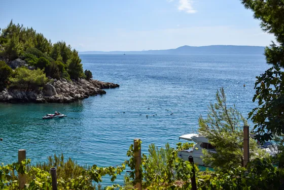 View of a clear blue bay with a small boat and yacht, rocky coastline, and lush greenery from a vineyard overlooking the Adriatic Sea near Split, Croatia.