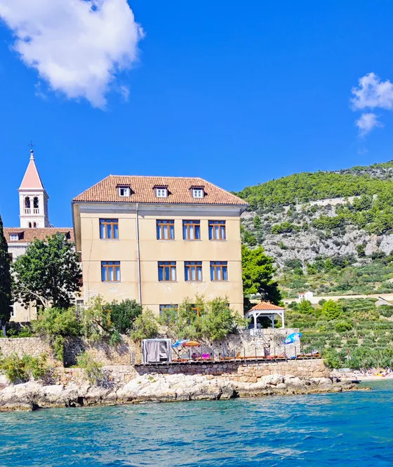 Historic Franciscan Monastery and bell tower on Hvar island's rocky coast, viewed from the turquoise Adriatic Sea.