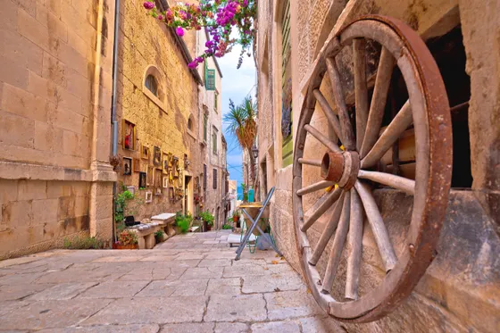 Narrow cobblestone alley in Korčula old town with antique wooden cartwheel, framed paintings and pink bougainvillea overhead