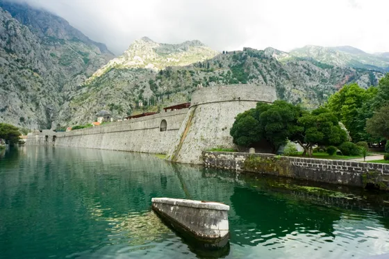 Massive medieval city walls of Kotor reflected in still green moat water with karst mountains behind