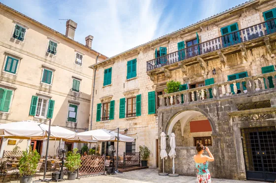 Tourist photographing historic stone buildings and green shutters of Kotor old town square, Montenegro
