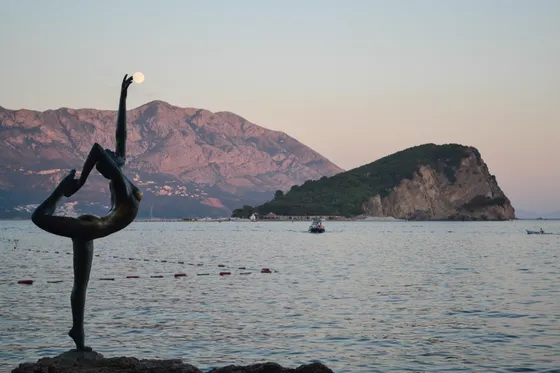 Budva's Ballerina (Dancer of Budva) bronze statue on the rocks at dusk with the full moon over Lovćen mountain and Sveti Nikola island in the background, Montenegro