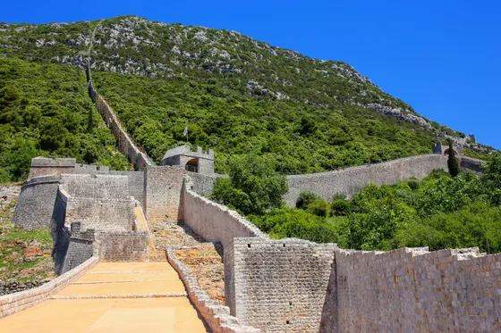 Ston defensive walls zigzagging steeply up a rocky green hillside with worn stone walkway in the foreground, Croatia